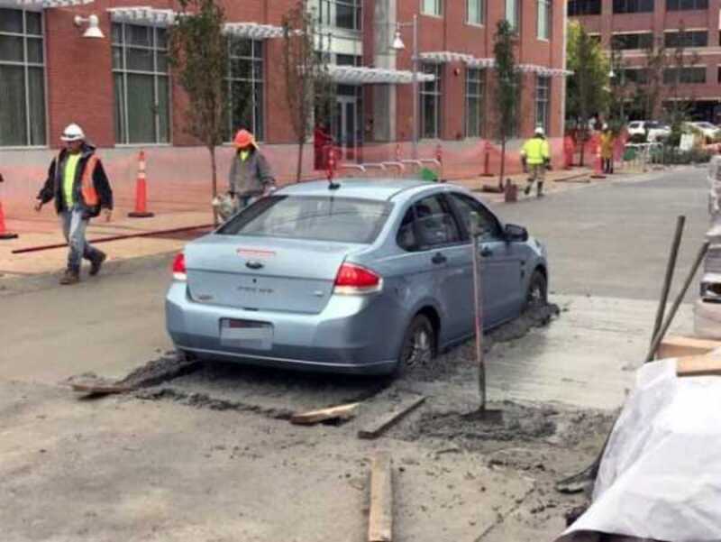 Car stuck in wet concrete with construction workers nearby.