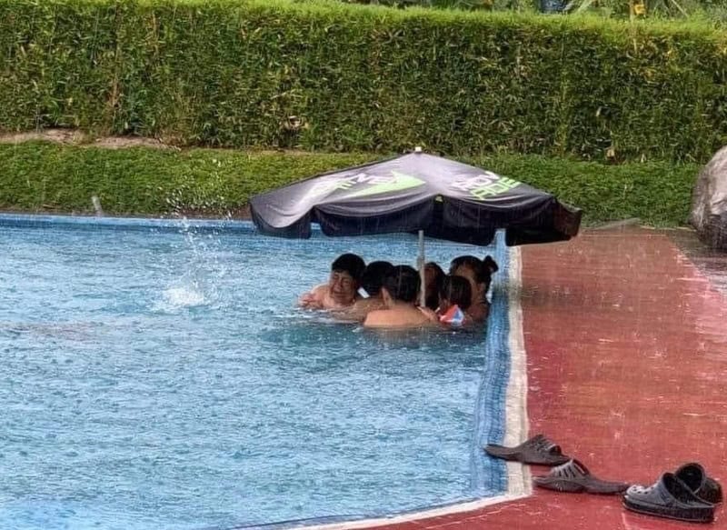 Group of people under a black umbrella in a swimming pool.