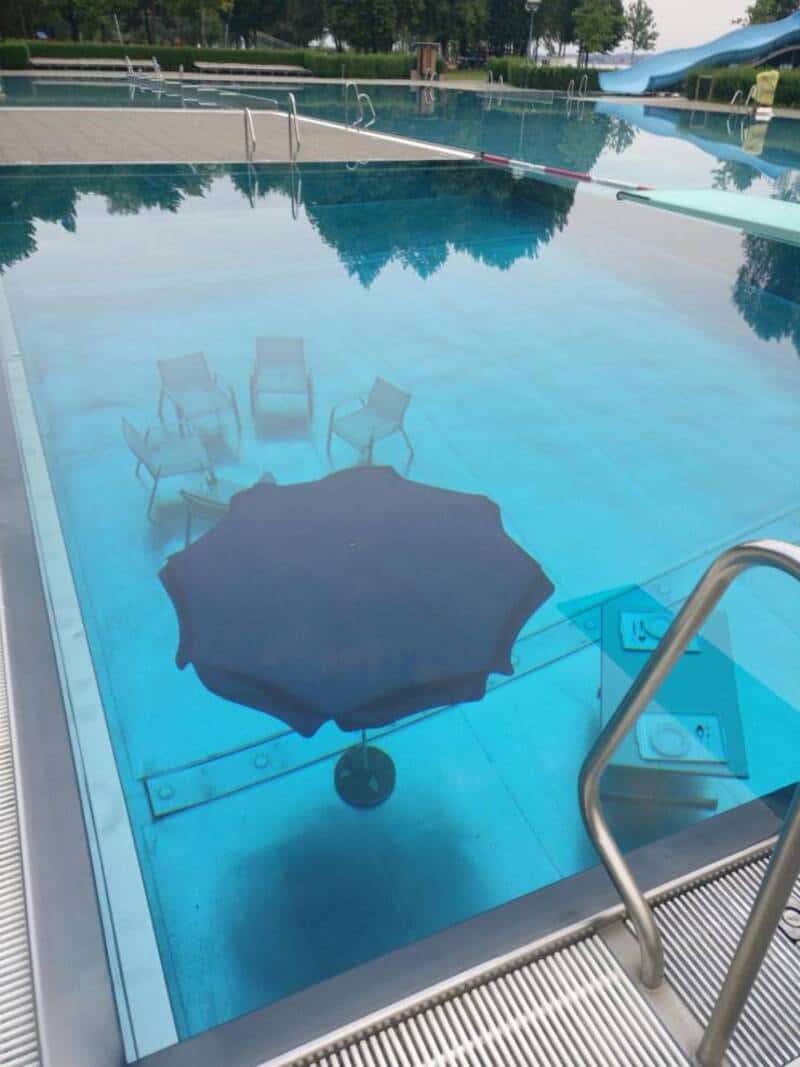 Umbrella submerged in a swimming pool with chairs and water slide in background.