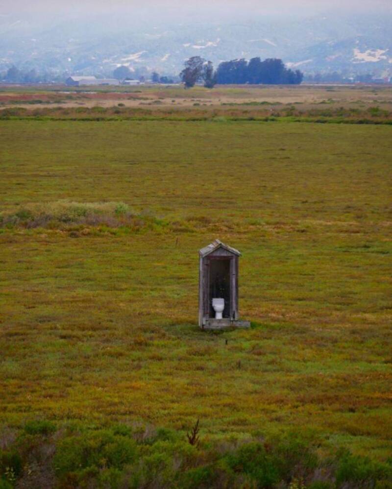 A portable toilet in a vast open field with mountains in the background.