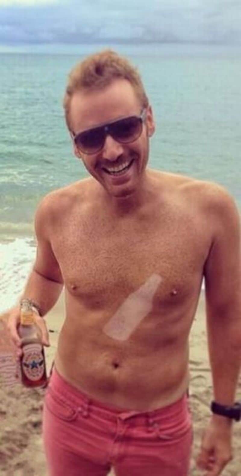 Man with sunglasses smiling on the beach holding a beer.