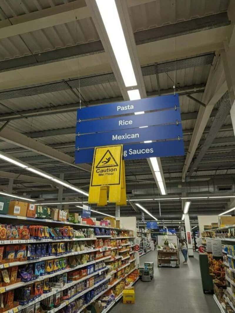 Sign in grocery store listing pasta, rice, Mexican, and sauces with caution sign below.