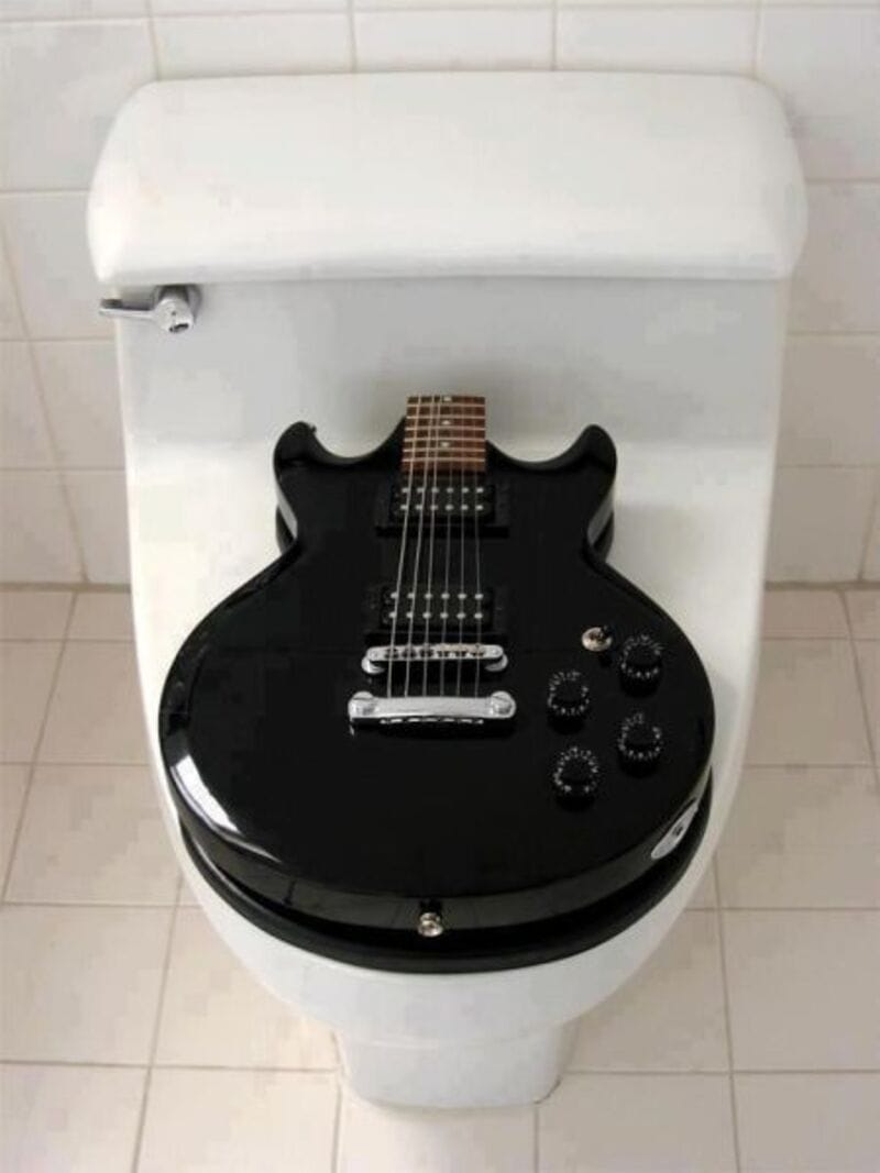 A black guitar placed on a white toilet seat lid.