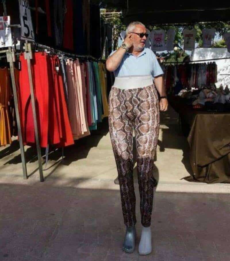 Man with snake-patterned pants and two different shoes at an outdoor market.