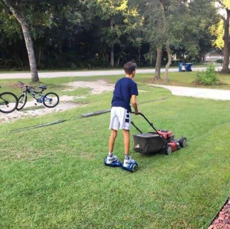 Boy mowing the lawn with a toy lawnmower on a scooter.