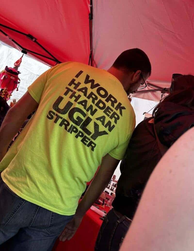 Man wearing a yellow T-shirt with the text "I WORK HARDER THAN AN UGLY STRIPPER" at an outdoor event.