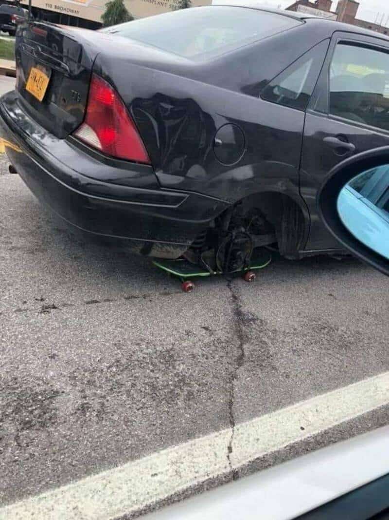 Damaged black car with flat tire and skateboard under rear wheel.