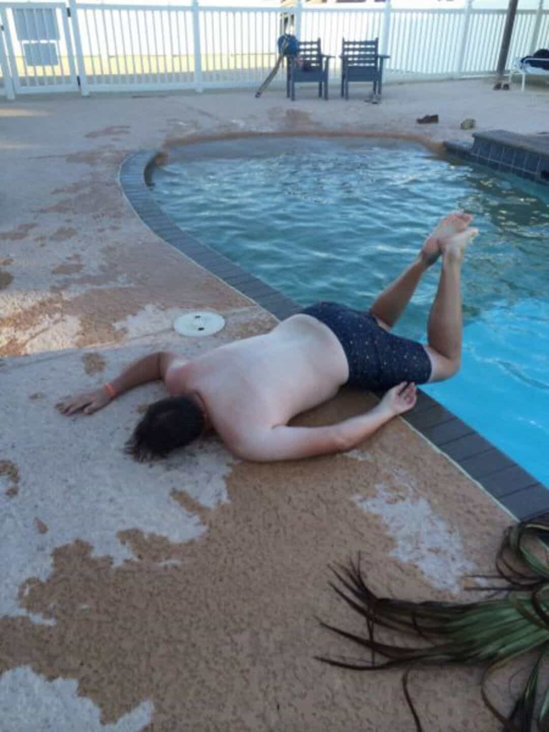 Person lying on pool deck with head near water, arm raised, wearing black swim trunks.