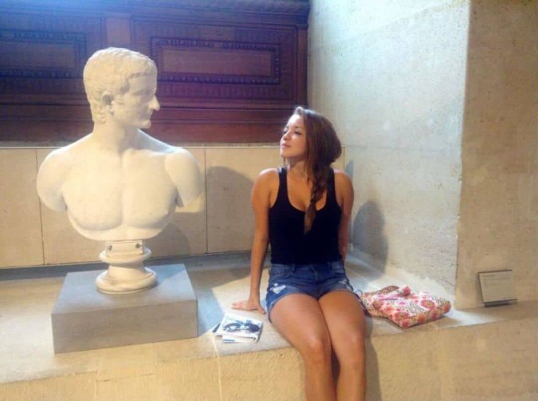 Woman sitting next to a bust sculpture in a museum, looking at it.
