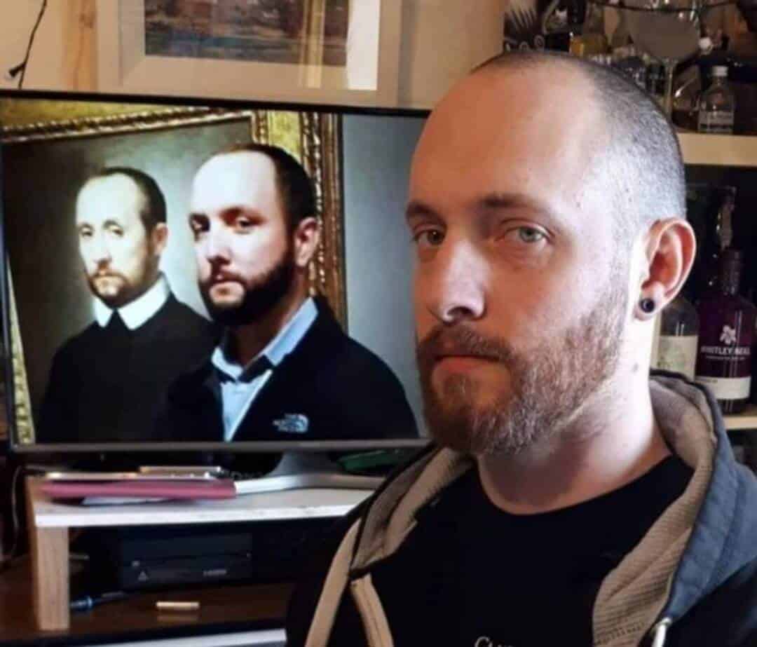 Man with beard and earring in front of a TV showing a portrait of two men.