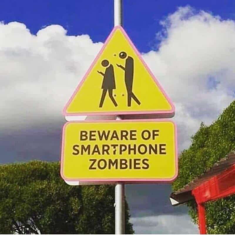 Warning sign warns of smartphone zombies with silhouettes of people looking at phones.