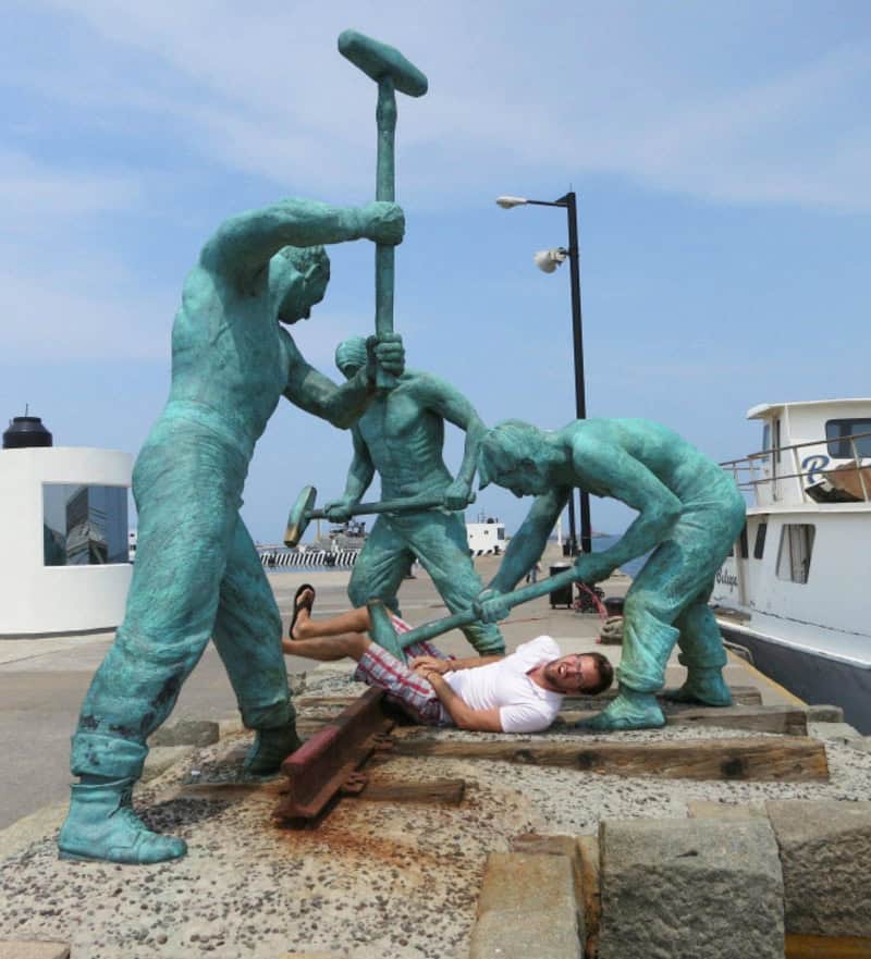 Statue of three men fighting with a woman lying on the ground at a harbor.