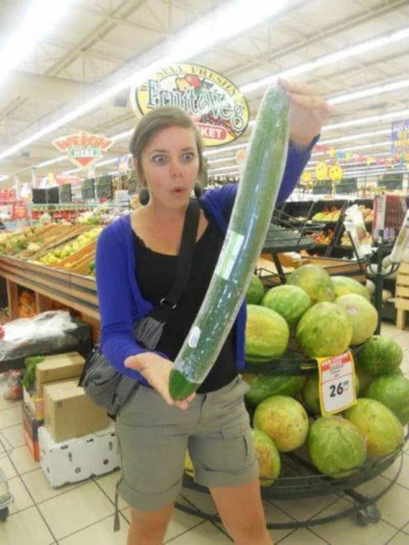 Woman holding a giant cucumber in a grocery store, surprised expression.