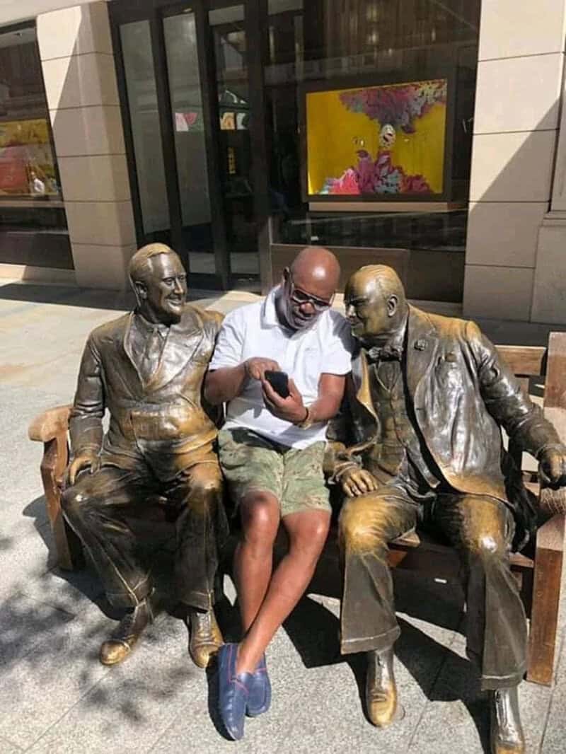 Man sitting between two bronze statues on a bench, looking at his phone.