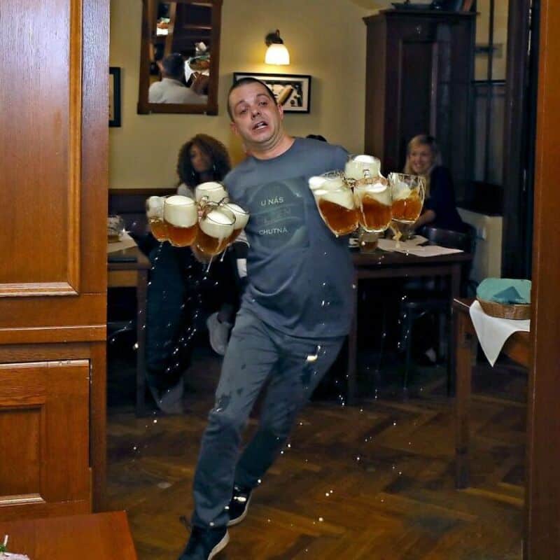 Man holding multiple beer glasses in a pub, with people in the background.