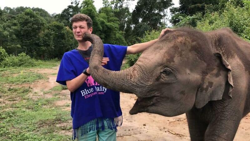 Young man with an elephant's trunk on his face in a green outdoor setting.