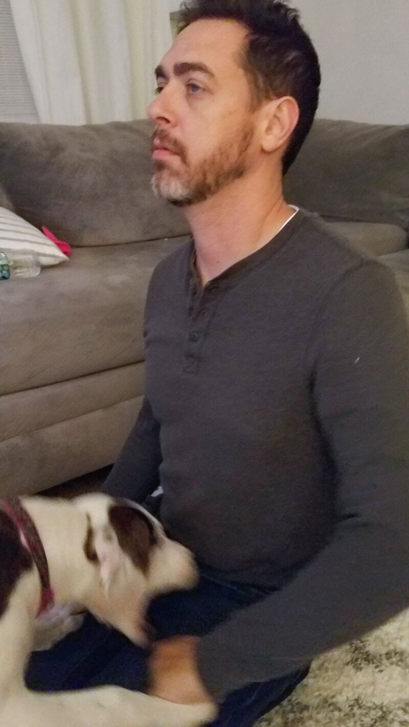 Man sitting on the floor with a dog, looking to the side, in a living room.
