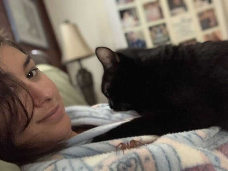Woman with dark hair lying on bed with black cat touching her face.