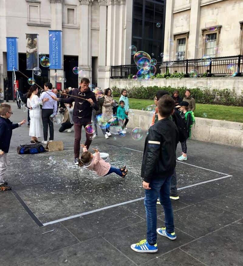 Children playing with bubbles outside a building, some falling and others floating.