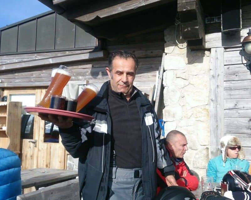 Man holding a tray with three glasses of beer outside a wooden building.