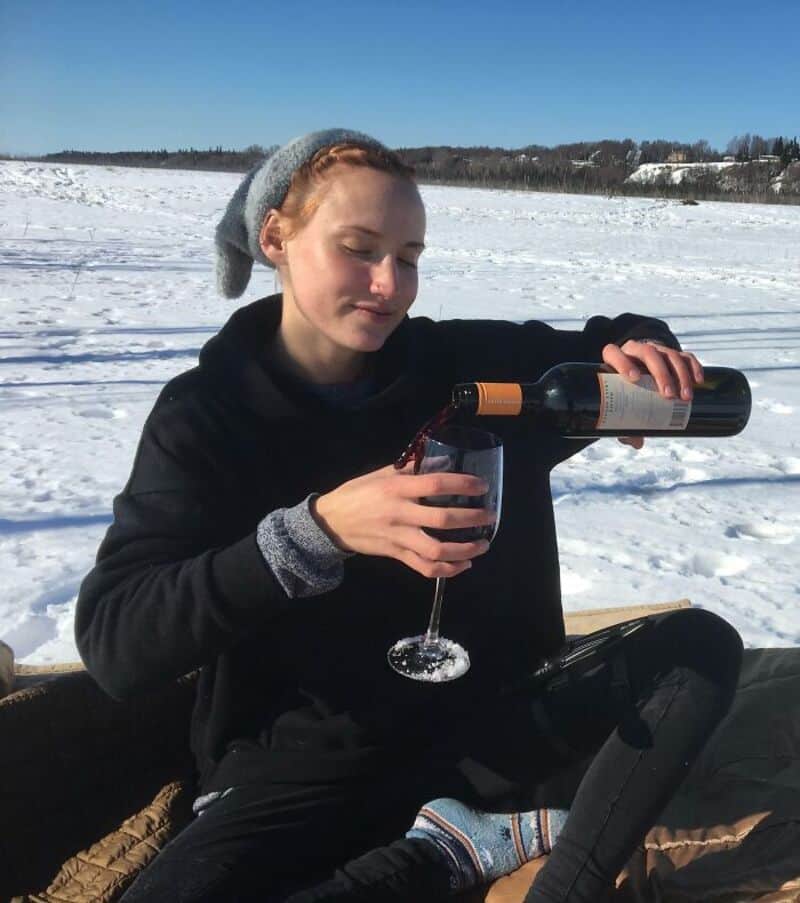 Young woman in winter clothes pouring wine in snowy landscape.