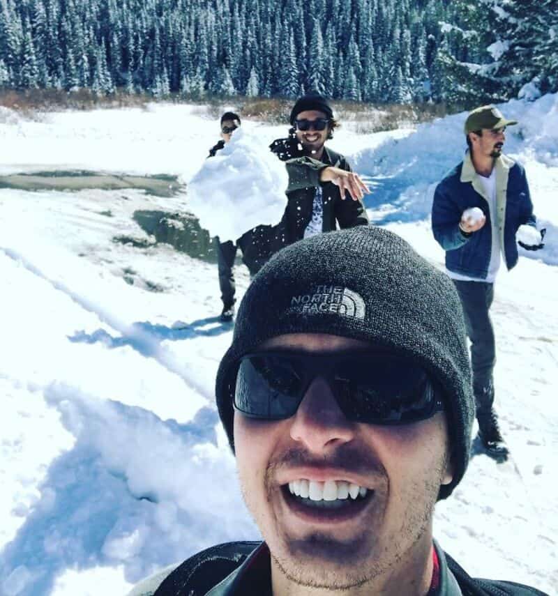 Group of friends enjoying snow, one taking a selfie, others playing with snowballs.