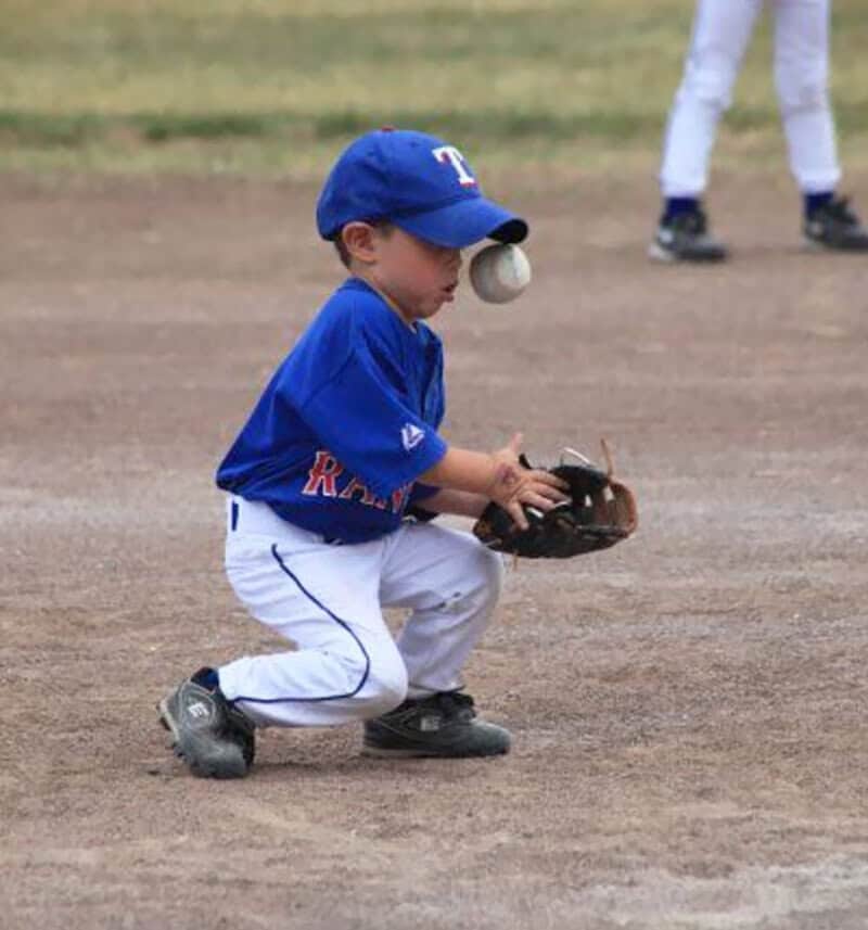 Young boy in baseball uniform catching a ball with his glove on the ground.