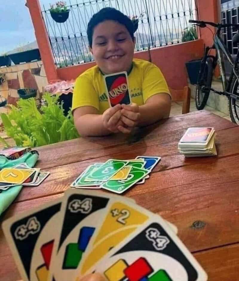 Child smiling holding UNO card game, playing at outdoor table with cards and deck.