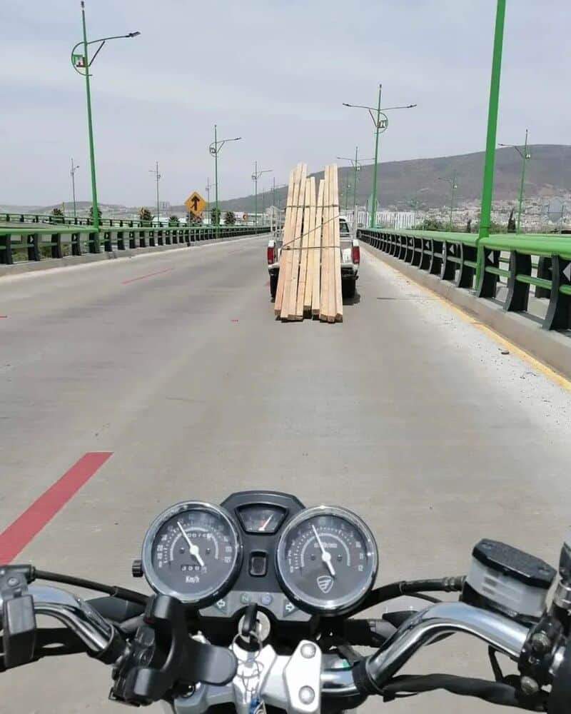 Motorcycle dashboard view of a bridge with wooden planks loaded on a vehicle ahead.
