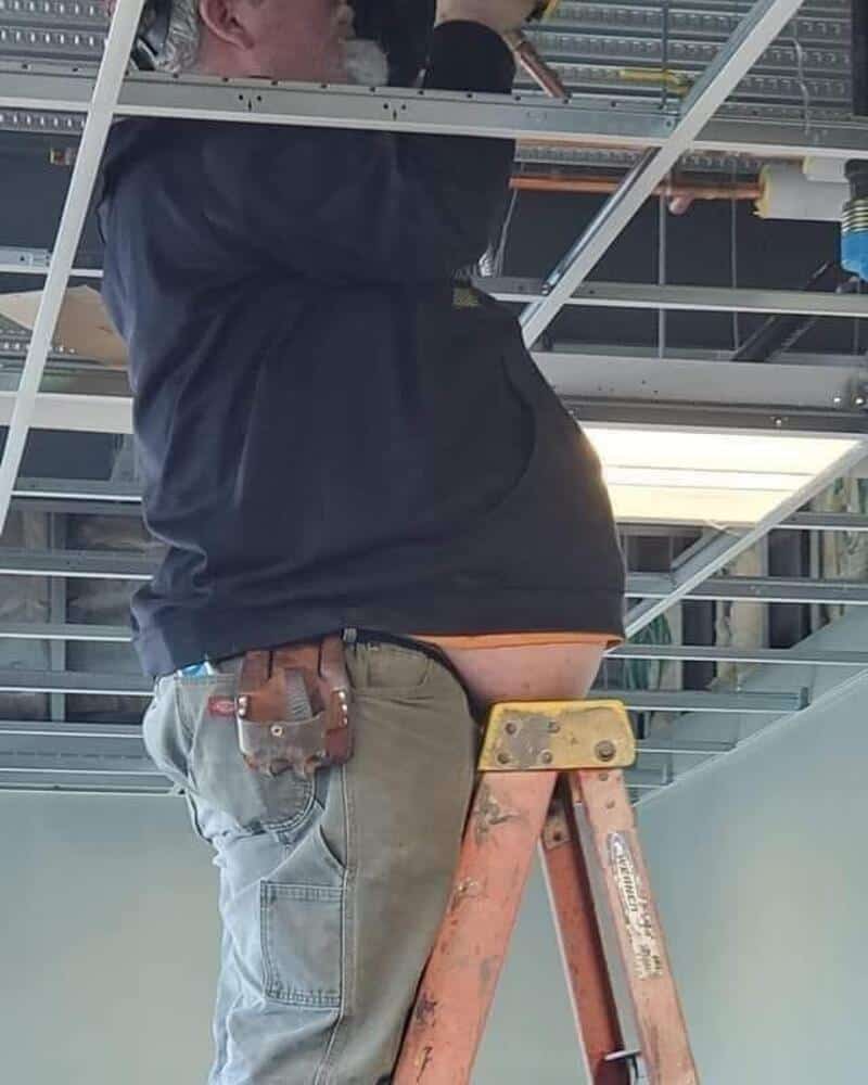 Person on ladder working on ceiling with exposed wiring and pipes.
