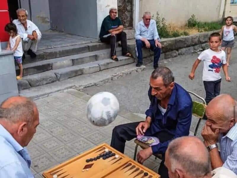 Men playing backgammon on the street with children watching.
