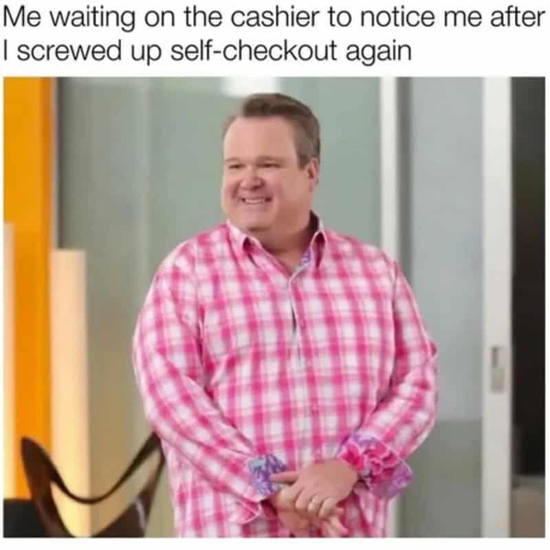 Man smiling in pink checkered shirt waiting at self-checkout.