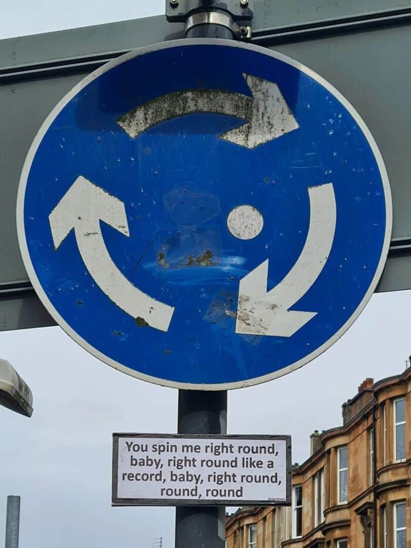 Roundabout sign with humorous sign below about spinning in circles.