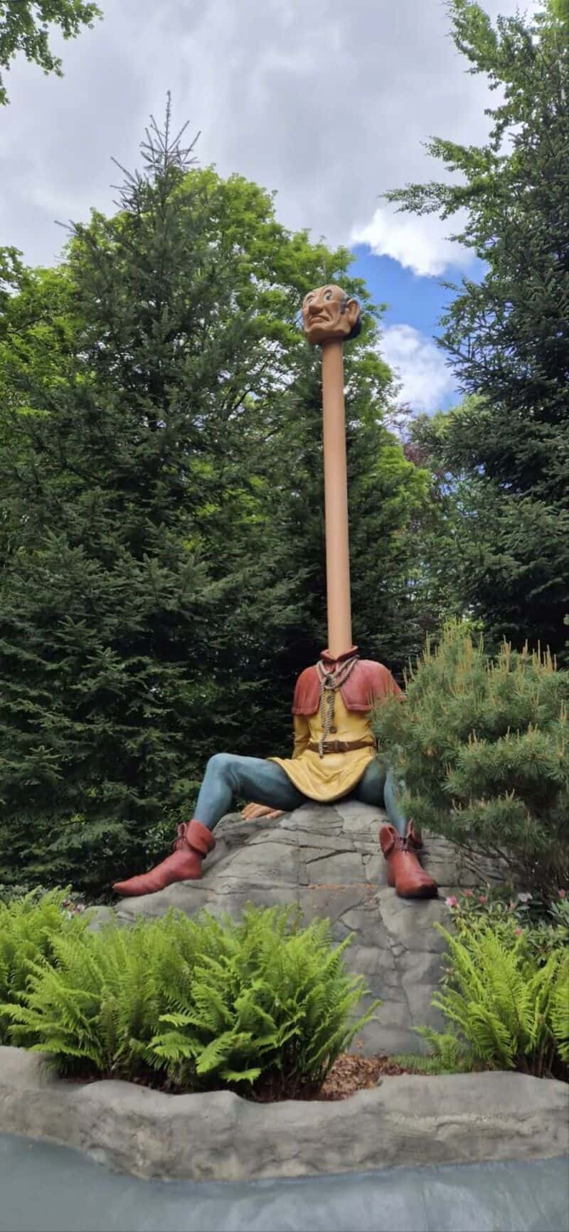 Sculpture of a person with a long neck and a head on a stick, sitting on a rock.
