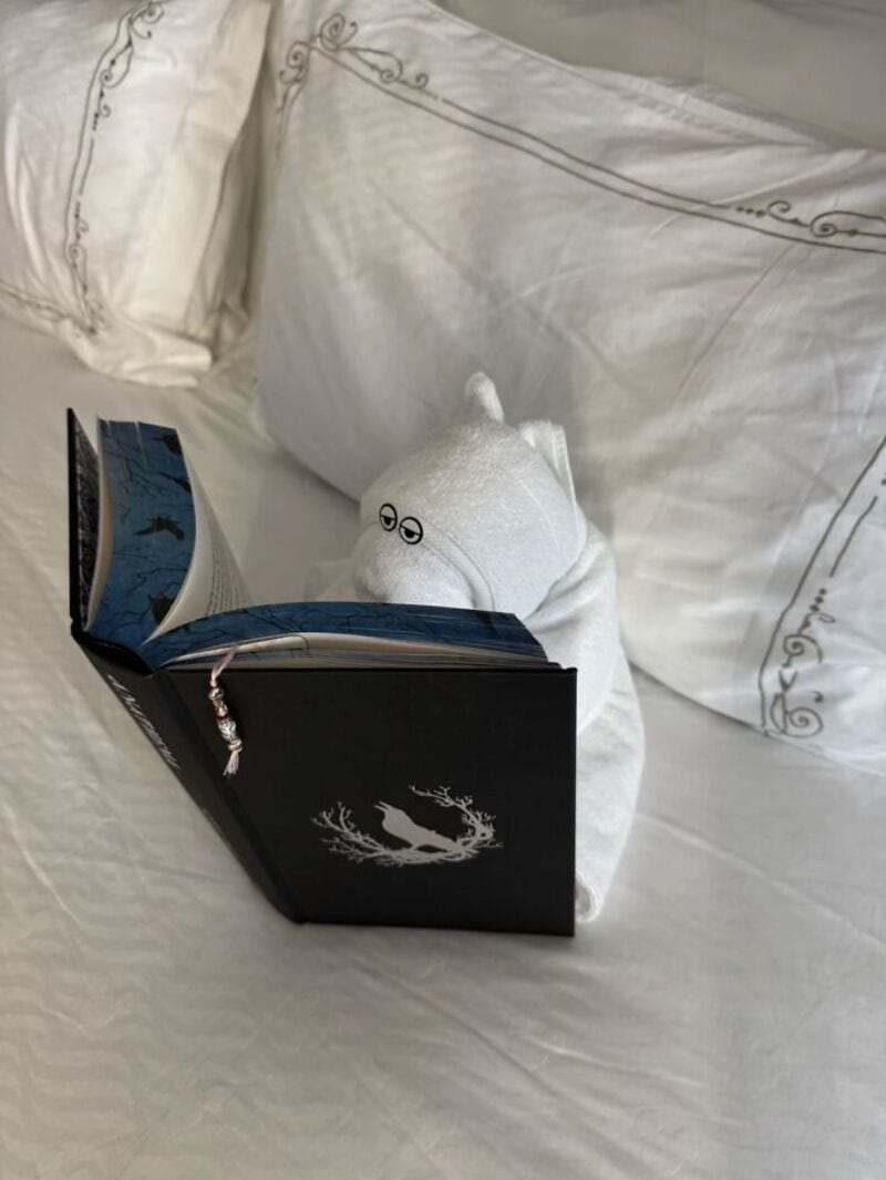 A white plush toy with glasses reading a black book on a bed.