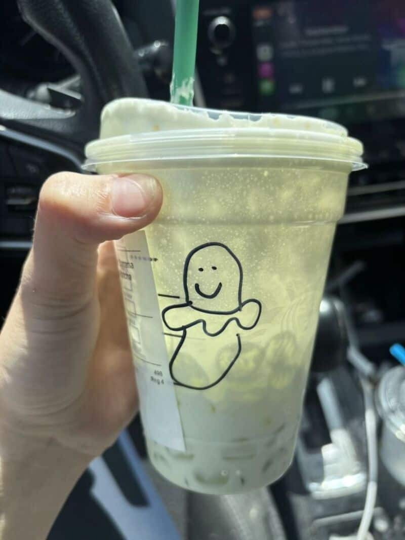 A hand holding a clear plastic cup with a green straw, featuring a drawing of a smiling ghost on the.