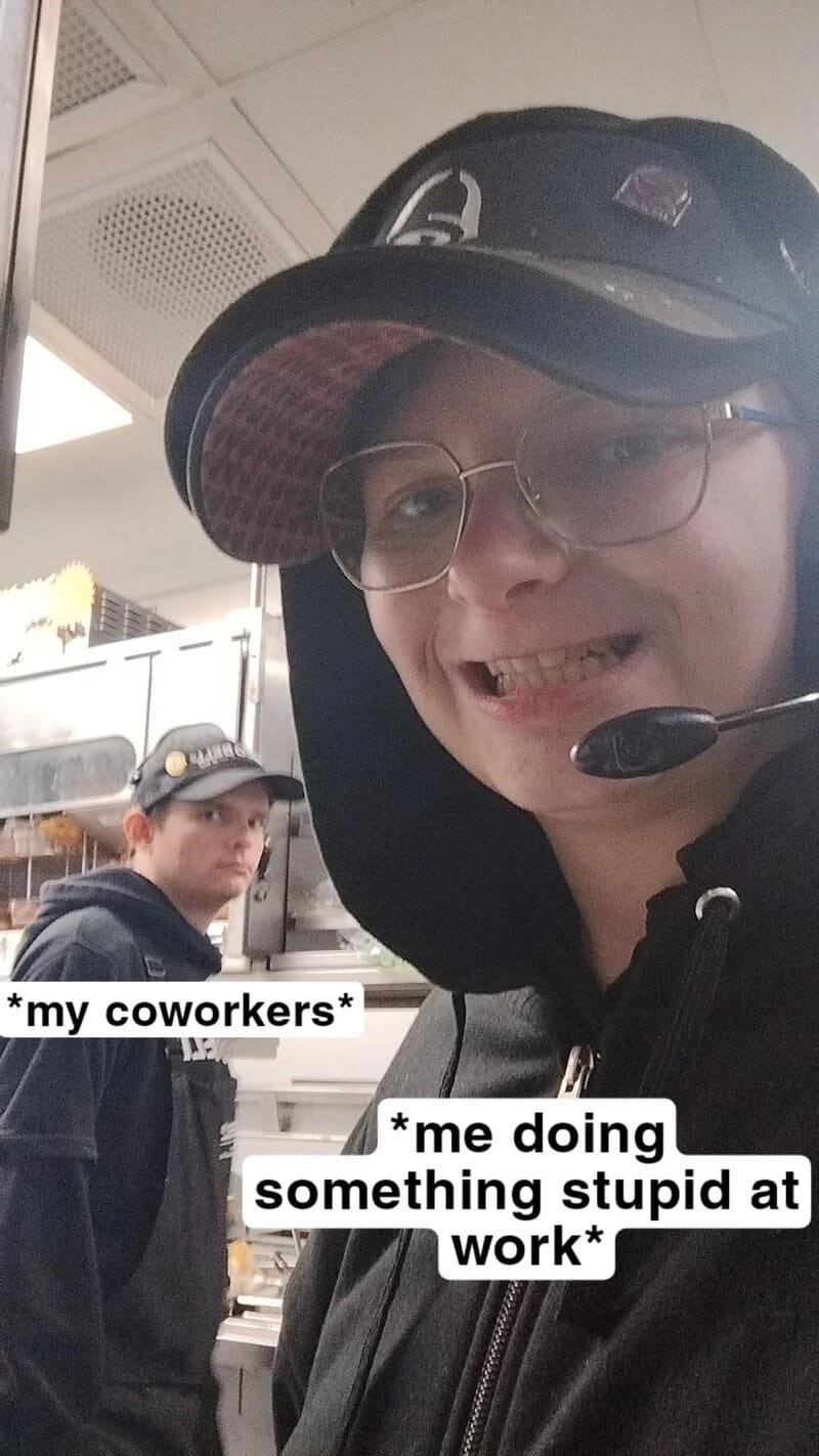 Person wearing glasses and a cap, smiling with a headset, in a work environment.