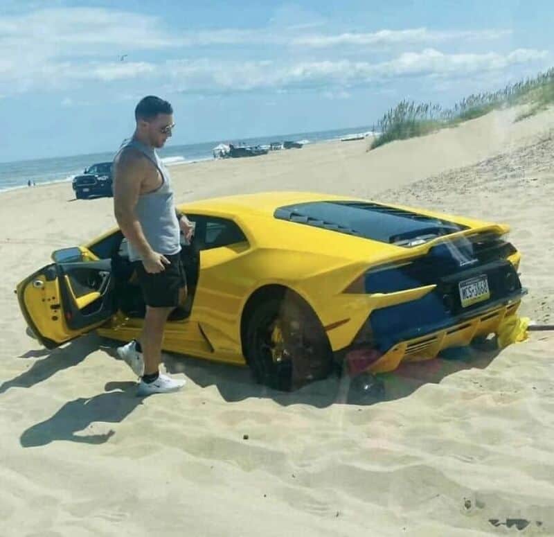 Yellow sports car stuck in sand with a man standing beside it on the beach.
