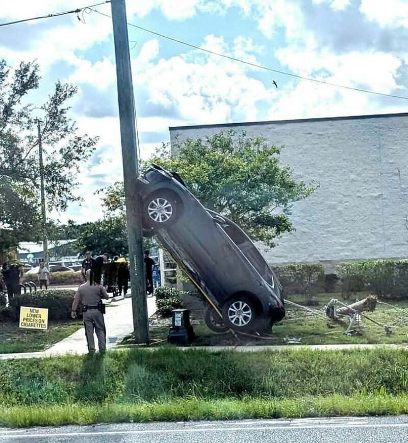 Car crashed into a pole, hanging at an angle with people observing.