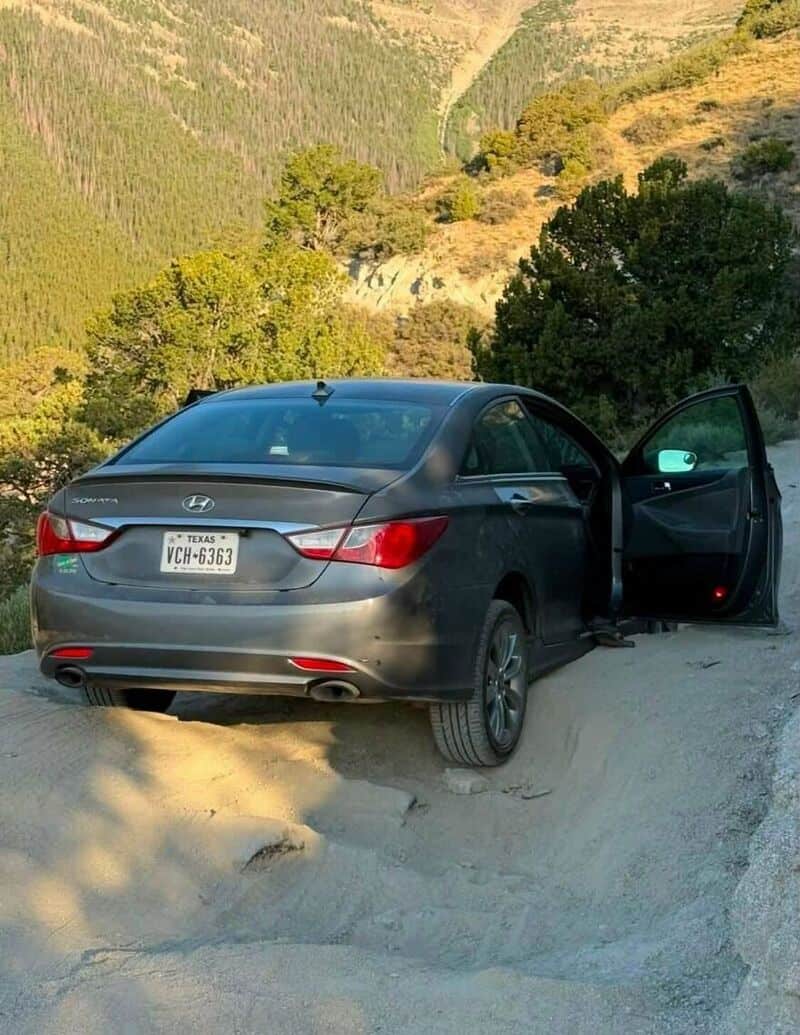 Car parked on a dirt road with open door, mountains and trees in background.