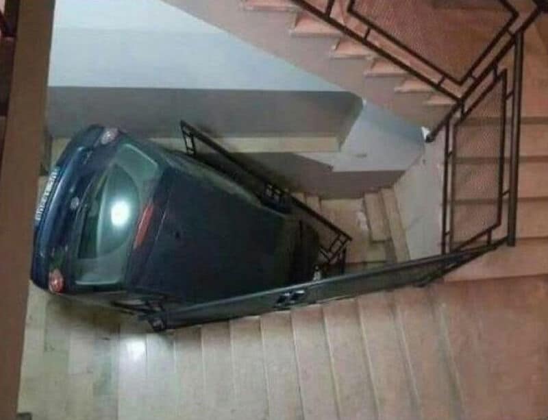 Car parked on staircase, blocking the way, with stairs and railing visible.