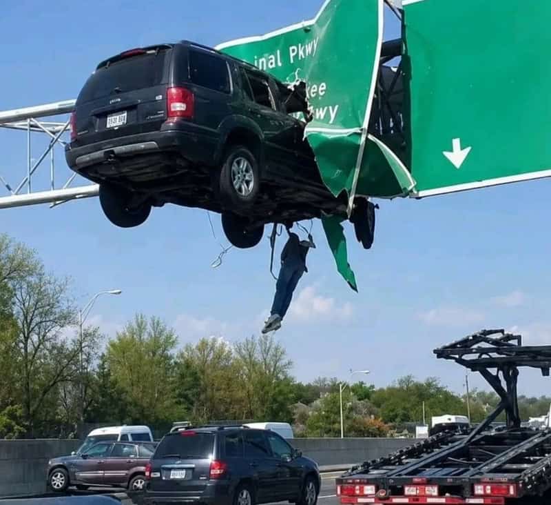 Car crashed into highway sign, hanging upside down with a person hanging from it.
