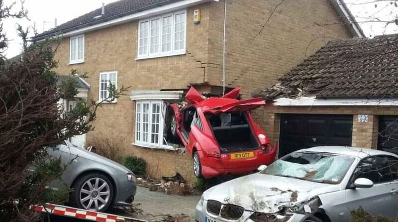 Red car crashed into house, hanging over the sidewalk, with other damaged cars nearby.