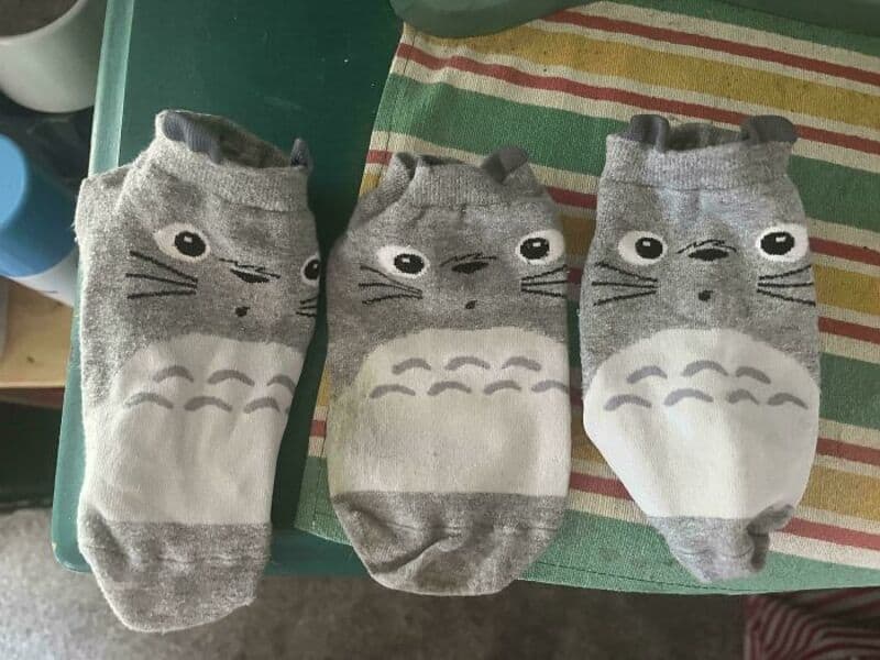 Three gray cat-themed socks with whiskers and eyes, laid on a striped cloth.