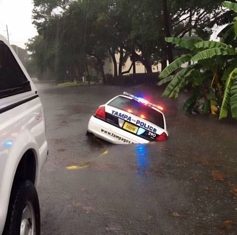 Flooded street with a partially submerged police car and a white vehicle nearby.
