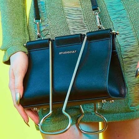 Black Studio Cult handbag with metal hooks and a strap, held by a person in colorful clothing.