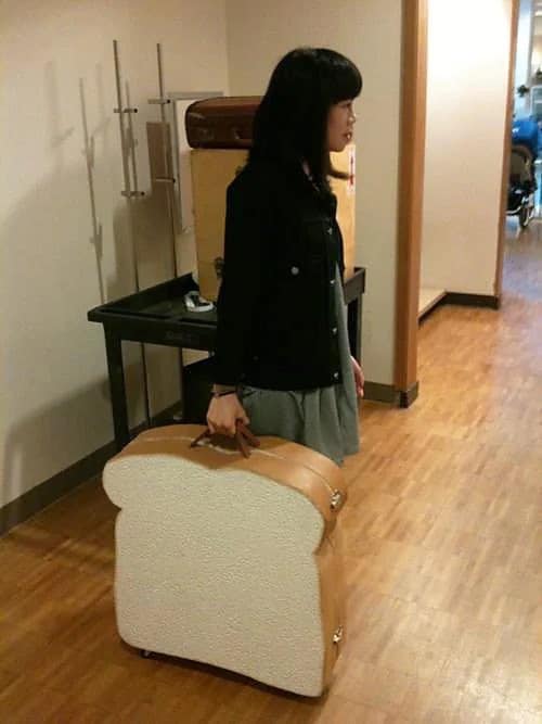 Girl with black hair in black jacket holding a suitcase shaped like a slice of bread.