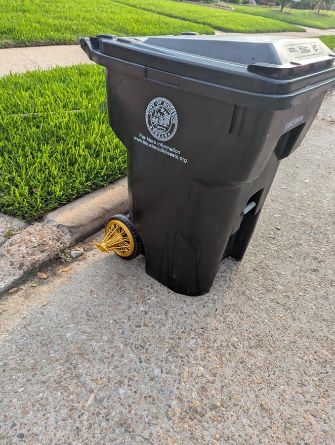 Black trash bin on street with yellow wheel and city logo.