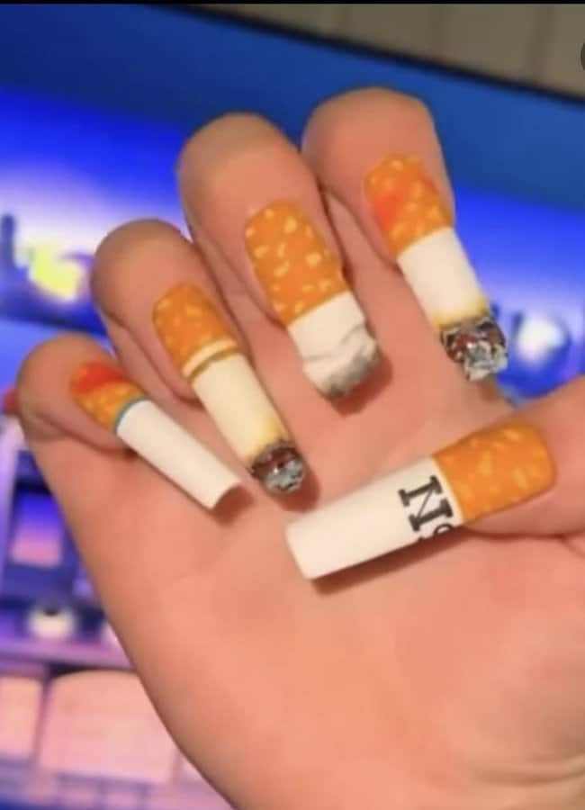 Fingers with cigarette-shaped nail art, some with fake diamonds, in front of a TV.