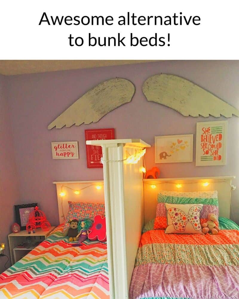 Two beds with colorful bedding separated by a white pillar, angel wings above, fairy lights, and art.